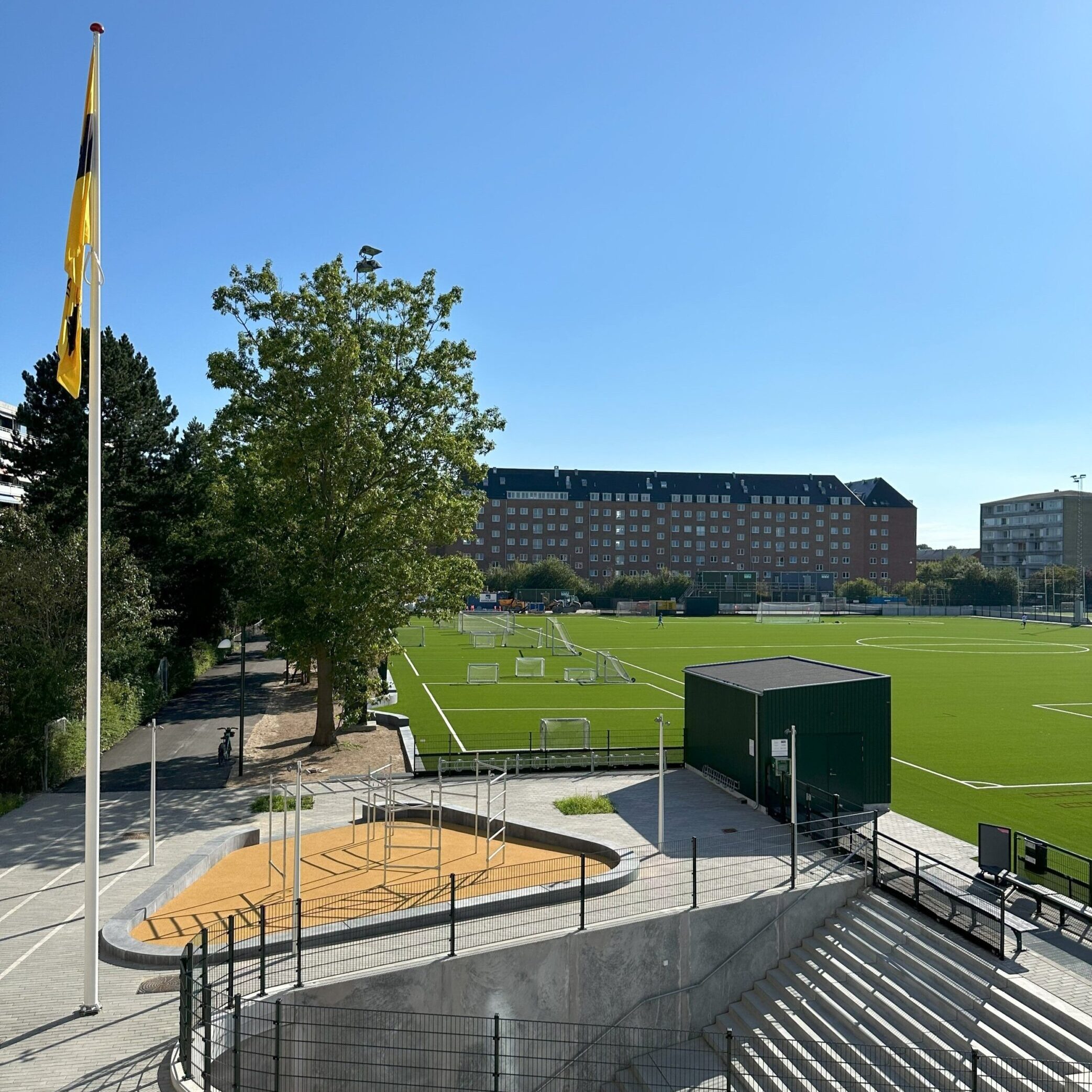Udsigt over Frederiksberg Boldklubs anlæg med grøn kunstgræsbane, tribune og flagstang med gult klubflag på en solrig dag.